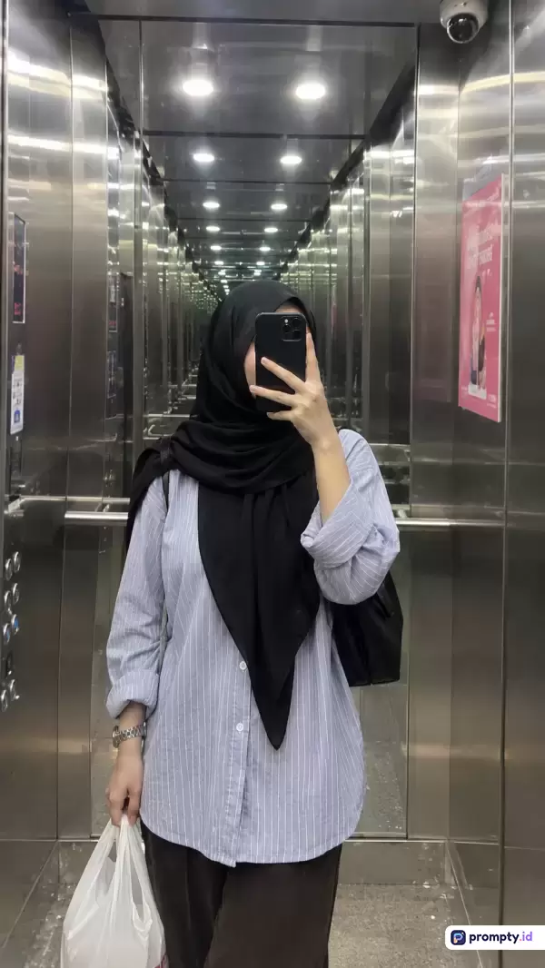 Gambar bergenre swafoto cermin bergaya modern ini menampilkan seorang wanita berhijab hitam dan kemeja garis-garis biru muda yang sedang berpose di dalam lift berdinding logam reflektif dengan dominasi warna perak, hitam, dan biru muda, serta kata kunci wanita, hijab, cermin, lift, swafoto.