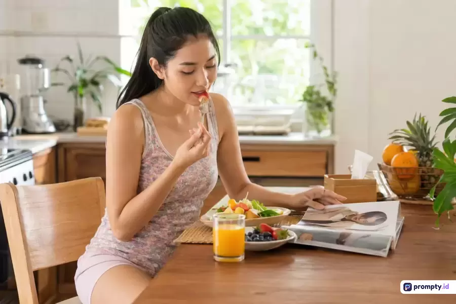 Foto candid bergaya gaya hidup sehat dengan pencahayaan alami menampilkan wanita Asia muda berambut hitam mengenakan pakaian santai bermotif bunga, sedang duduk di meja kayu dapur cerah sambil makan buah segar dan membaca majalah, dikelilingi warna dominan cokelat kayu, hijau tanaman, dan kuning jeruk segar dengan kata kunci wanita, sehat, makanan, dapur, gaya hidup, majalah, buah.