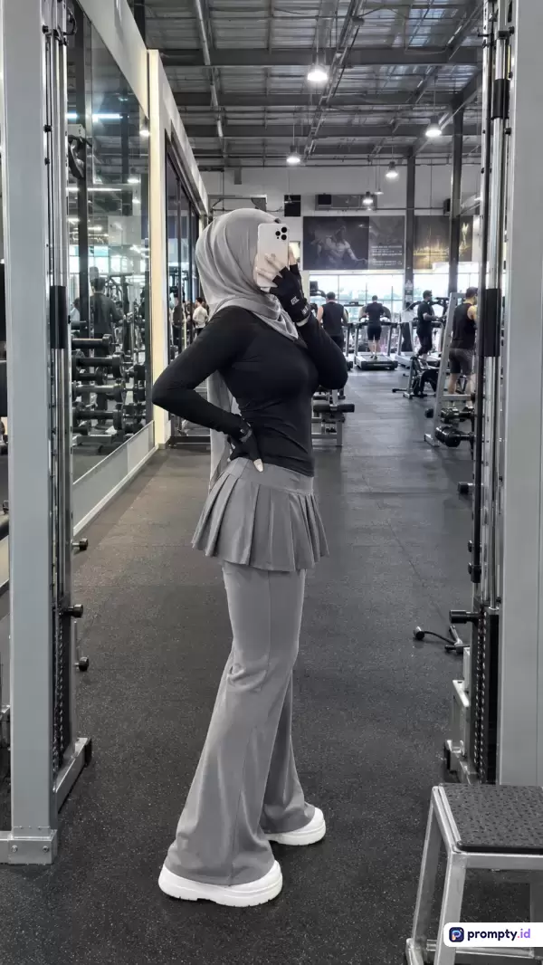 Gambar gaya fotografi selfie cermin ini menampilkan seorang wanita berhijab yang sedang berpose di dalam gym modern dengan gaya kasual-sporty, menggunakan kata kunci wanita berhijab, gym, fitness, olahraga, mirror selfie, dengan dominasi warna abu-abu, hitam, dan putih.