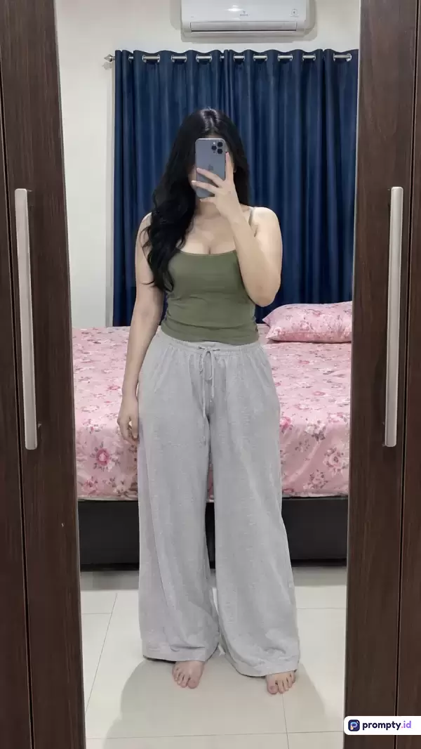 Gambar bergaya swafoto kasual ini menampilkan seorang wanita berambut hitam panjang yang berdiri di depan cermin kamar tidur mengenakan tank top hijau zaitun dan celana panjang longgar abu-abu, dengan latar belakang tirai biru tua dan tempat tidur bermotif bunga, menghasilkan suasana santai dan intim yang diperkuat oleh dominasi warna hijau zaitun, abu-abu, dan biru tua, serta kata kunci swafoto cermin, pakaian santai, kamar tidur, gaya kasual, dan fesyen rumah.